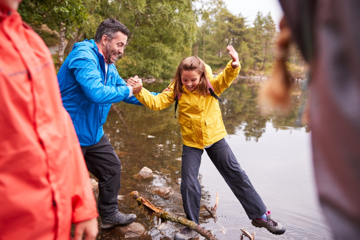 Family enjoying a walk in the Lake District by the side of a lake. A young girl is walking on the rocks at the edge of the lake with her parents, surround by a trees in a forest.